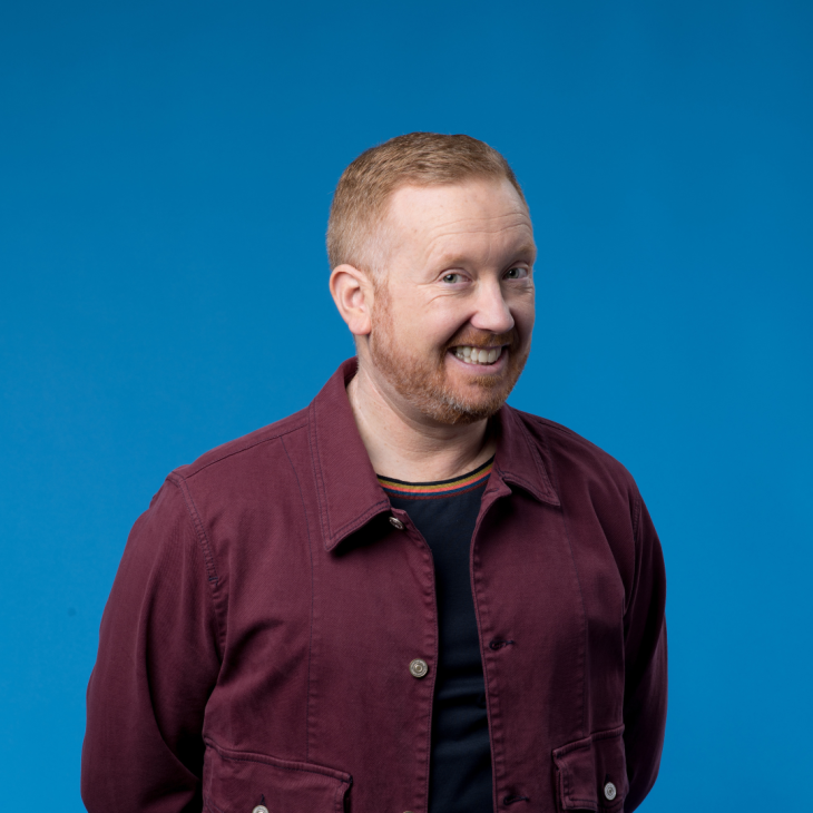 Luke McGregor has short red hair and a beard. He is standing in front of a blue background and is wearing a t-shirt with a maroon button up over the top. He is smiling with his teeth and looking into the camera.