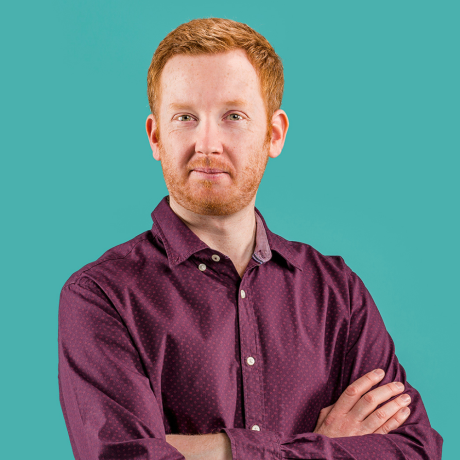 Luke has his arms crossed and is turned slightly his left. His head and gaze are turned directly to camera. Luke has short red hair and a short-trimmed beard & moe. His lips are pursed and smiling slightly. Luke wears a maroon long-sleeved button up shirt with a feint, light pattern. The background is a teal green.