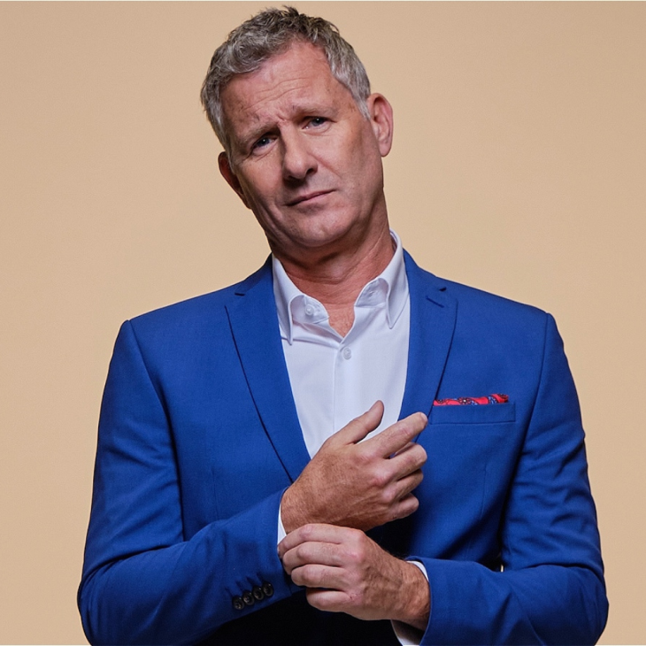 Adam has short grey hair. He is wearing a blue suit, blue shirt and re pocket square. He is looking into the camera and fixing his cuff.