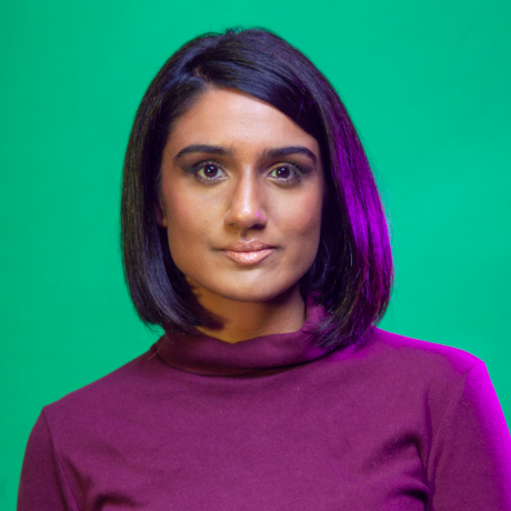 Vidya Rajan looks straight to camera with a neutral expression. She has deep, brown eyes. Her gently curved black, shoulder length hair frames her face. She is wearing a purple skivvy, which off sets the purple light reflecting off her hair to her left. The backdrop is mid-green.