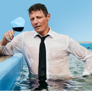 Image is of Merrick Watts wearing a white dress shirt with a black tie. He is holding a glass of red wine and standing in a pool.