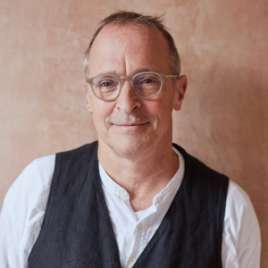 David has wispy mid-brown hair with greying side burns. His eye glasses have light brown frames and he is smiling warmly at the camera. He has a white shirt with the top two buttons undone, under a charcoal grey vest. He is front of a textured, caramel coloured background.