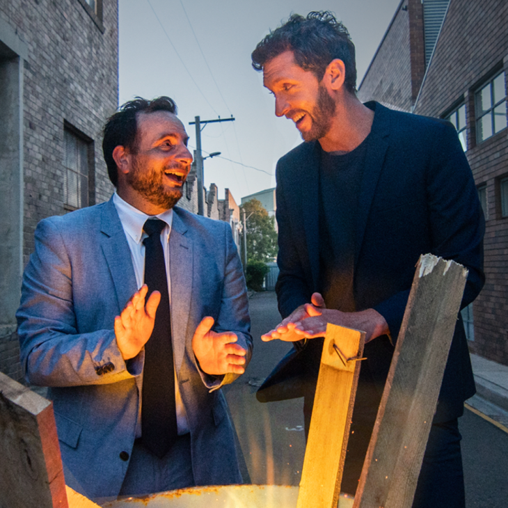 Dan Ilic and Lewis Hobba are standing in a back street, smiling at each other as they warm their hands on a fire which is burning inside a petrol drum. There are a few different lengths of wood sitting in the drum, poking up from the flames. The brick walls of the buildings on either side of the street run quite a way beyond where they stand. In the distance, in the gap between the walls, we can see blue sky. Dan is to the left of shot. He has short brown hair and pronounced stubble. He wears a light bluer suit, an even lighter blue business shirt and a chocolate brown tie. Lewis is to the right of shot. He has short brown hair and a trimmed beard & moe. He wears a midnight blue jacket over a t-shirt of the same colour.