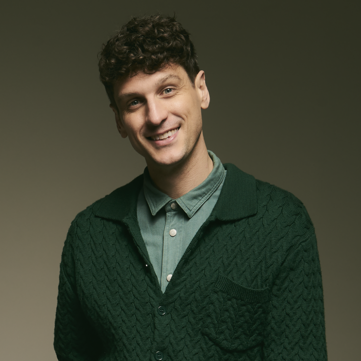 Image is of Steen Raskopoulos. Steen has short brown curly hair. He is wearing a light green button up shirt and dark green cardigan. He is standing in front of a dark green background and is smiling into the camera.