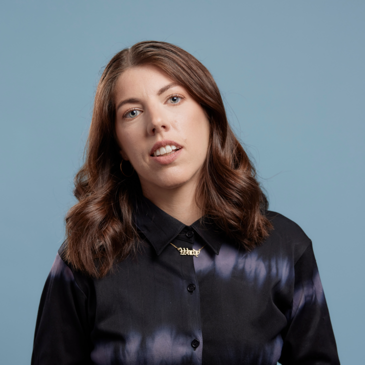 Alex Ward stands front on to the camera, her head tiled gently to her right. She smiles gently with her teeth showing and her blue eyes look directly and warmly to the camera. Her long brown hair drops below her shoulders. Her button up long sleeved shirt is black silk with the odd row of soft white tie die blurs. Between her collar ends is a fine gold chain with the word Wardy in stately lettering. The background is a grey blue.