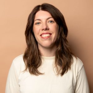 Image is of Alex Ward. Alex has medium length brown hair and is wearing a off-white sweater. She is standing in front of a light brown backdrop and smiling.