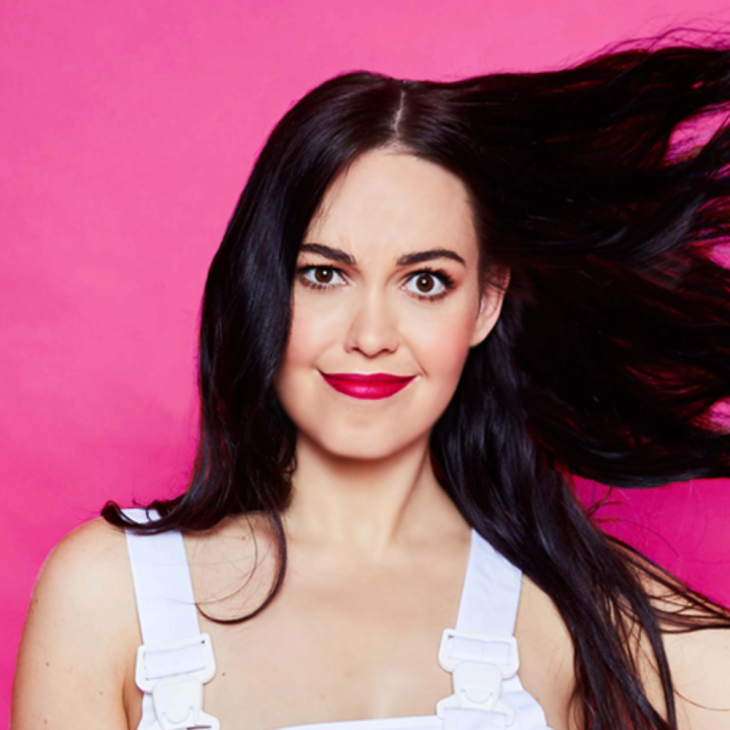 Bec has long black hair which is being blown to her left. She has bright red lipstick on and a mischievous, crooked smile. Her dark brown eyes are looking straight into the camera. She is in white overalls and the background is pink.