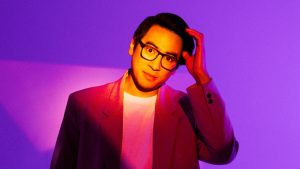 Image is of Michael Hing. Michael is wearing an orange suit jacket, white t-shirt and dark framed reading glasses. His left hand is raised to his left temple. The lighting is effecting this image immensely and creating different shades of purple.