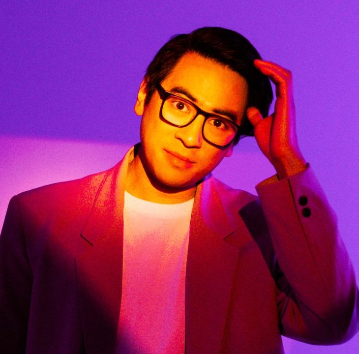 Image is of Michael Hing. Michael is wearing an orange suit jacket, white t-shirt and dark framed reading glasses. His left hand is raised to his left temple. The lighting is effecting this image immensely and creating different shades of purple.