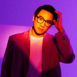 Image is of Michael Hing. Michael is wearing an orange suit jacket, white t-shirt and dark framed reading glasses. His left hand is raised to his left temple. The lighting is effecting this image immensely and creating different shades of purple.