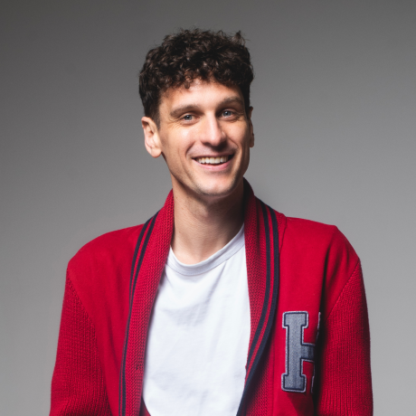 Steen Raskopoulos smiles welcomingly to camera, his head turned slightly to his left, his gaze direct into the lens. He has curly dark brown hair with short sides, blue eyes and he is clean shaven. He wears a white t shirt under a red cardigan with dark blue trim around the collar and continuing down the open front, with a light grey H on the left breast. The cardigan is much like the 1960’s fraternity house ones. The background is mid-grey.