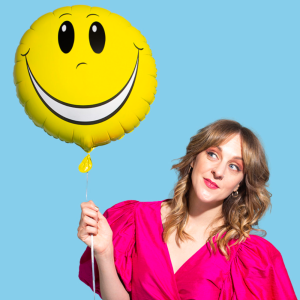 Gillian has long blonde curly hair. She is wearing a bright pink top and holding a yellow balloon with a smiley face. She has pink eye shadow on and a gloss on her lips. The background is light blue.