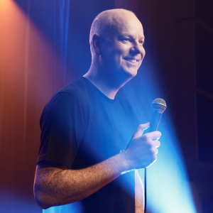 Image is of Tom Gleeson standing on stage holding a microphone. A stage light is shooting up from the bottom right corner, directly hitting his face. Tom is wearing a black t shirt and smiling out of frame.