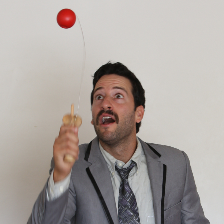 Ben is wearing a dark grey suit, light grey top and dark grey tie. He is bouncing a red paddle ball and looking up at it. He has an excited expression on his face. The photo has been taken in a lounge room.