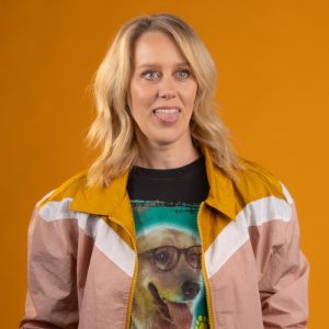Claire has short blonde wavy hair. She is wearing a colourful jacket that is yellow, white and pink over a graphic t-shirt. She is looking off camera and poking out her tongue. The background is a mustard colour.