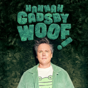 Image is of Hannah Gadsby. Hannah is wearing a green corduroy jacket and a white t-shirt with a picture of a cartoon frog on it. They have short grey hair and green eyes. They are standing in front of a green backdrop that has a fluffy texture.