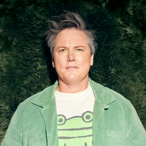 Image is of Hannah Gadsby. Hannah is wearing a green corduroy jacket and a white t-shirt with a picture of a cartoon frog on it. They have short grey hair and green eyes. They are standing in front of a green backdrop that has a fluffy texture.