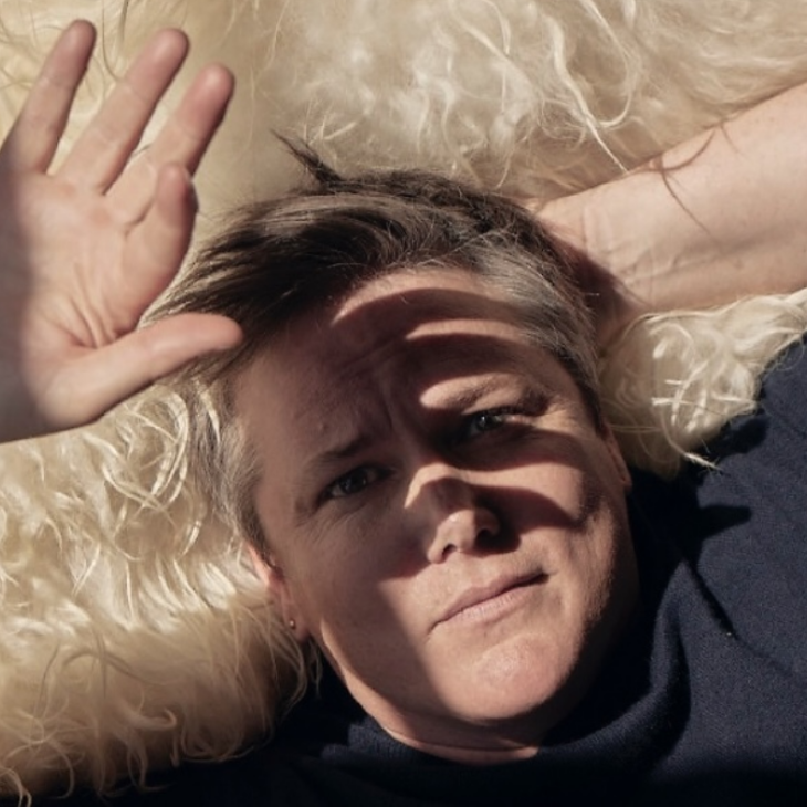 Comedian Hannah Gadsby laying down on a rug, shading their face with their hand.