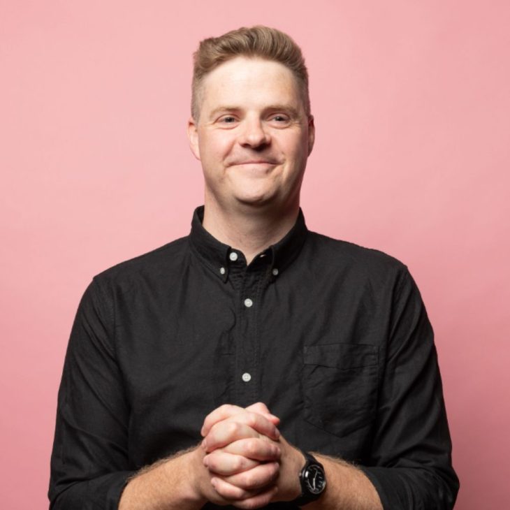 Tom Ballard has short blonde hair and blue eyes. He is wearing a black buttoned up top and is standing in front of a light pink backdrop. He is smiling just outside of the camera lens and has his hands clasped together in front of his stomach.