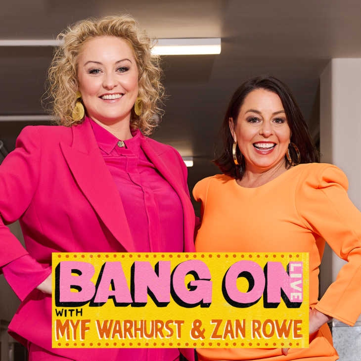 Image is of Zan Rowe (left) and Myf Warhusrt (right) smiling directly into the camera lens. Zan has shoulder length curly hair and is wearing a bright pink suit and gold dangly earrings. Myf is wearing a bright orange dress and gold hooped earrings.