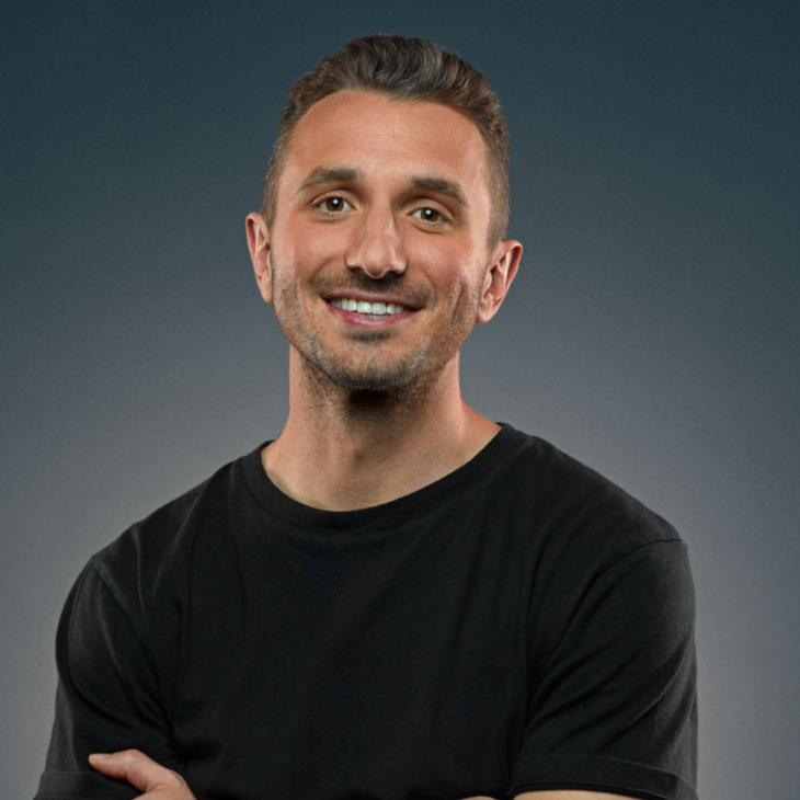 Tommy Little has brown hair, brown stubble and brown eyes. He is wearing a black tshirt and has his arms crossed. He is looking toward the camera and smiling. The background is a dark blue.