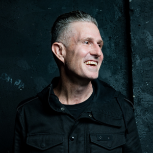 Wil Anderson has short grey hair. He is wearing a black shirt and jacket. He is looking to his right and smiling out of frame. The background is a textured black.