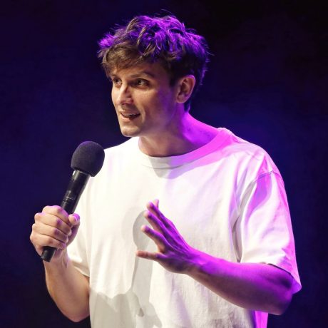 Sam Taunton is holding a microphone in the left hand and gesturing with the right hand. He is wearing a plain white t-shirt and is illuminated by purple stage lighting against a dark background.