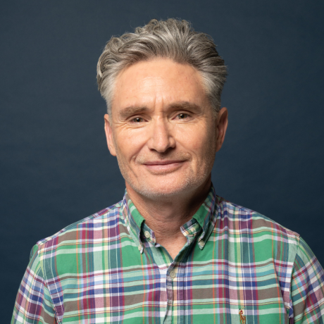 Image is of Dave Hughes. He is wearing a green, red & blue checked shirt. He is looking towards the camera. He has short wavy grey hair and stubble. The background is navy blue.