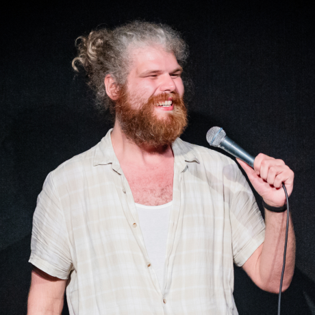 Dave Woodhead is on stage with a dark grey curtain behind him. He is smiling whole-heartedly with a microphone in his left hand raised and pointed towards his mouth. He is looking off camera, to his left. Dave has curly hair, which is mostly grey, but has white puffs at the front. His hair is tied back behind his head. He has a full, fairly bushy red beard and moustache. Hear has a loose short-sleeved shirt on which is a very pale green with a white checked pattern. The the top three buttons are undone, showing a white singlet underneath.