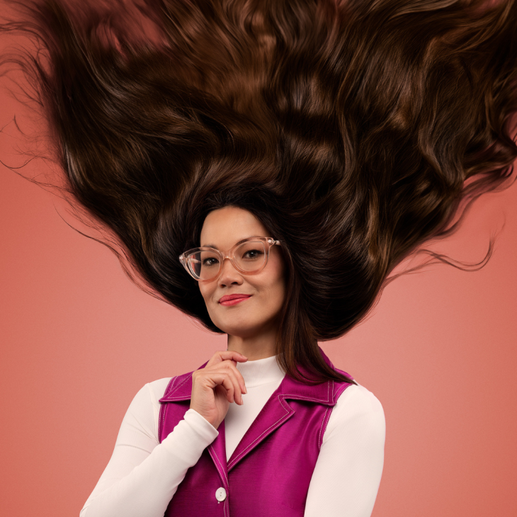 Lizzy Hoo smiles gently to the camera as her voluminous brown hair flies upwards in a V. She is turned slightly to her right. She wears a crisp white skivvy under a pink satin V neck topped dress with one large white button on it. She has large, translucent pink framed glasses on. Her right hand is up and almost closed, except for her index finger which sits above the others as if she is about to place it on her chin. The background is orange.