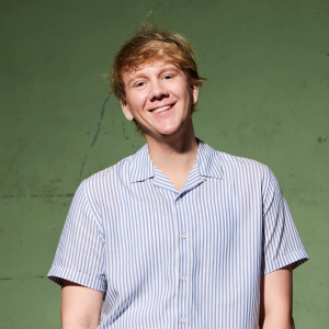 Josh is wearing a blue and white striped, short-sleeved shirt. His blonde hair is disheveled and his head is tilted slightly to the right as he smiles generously at the camera. The background is a concrete slab painted Brunswick green.