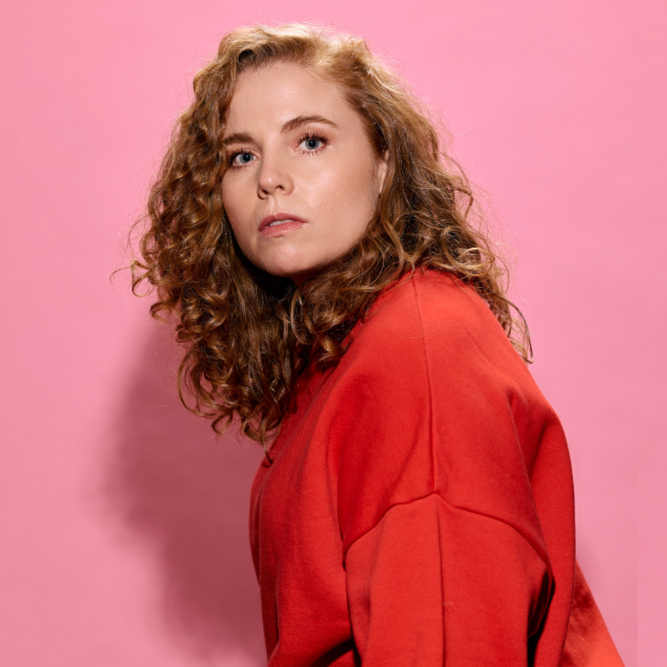 Bronwyn Kuss has her side to the camera, facing to her right, with her head turned to the camera. Her long, curly, strawberry blonde hair cascades over her shoulder. Her expression is quite neutral. She wears an orange jumper. She casts a shadow onto the baby pink background behind her.