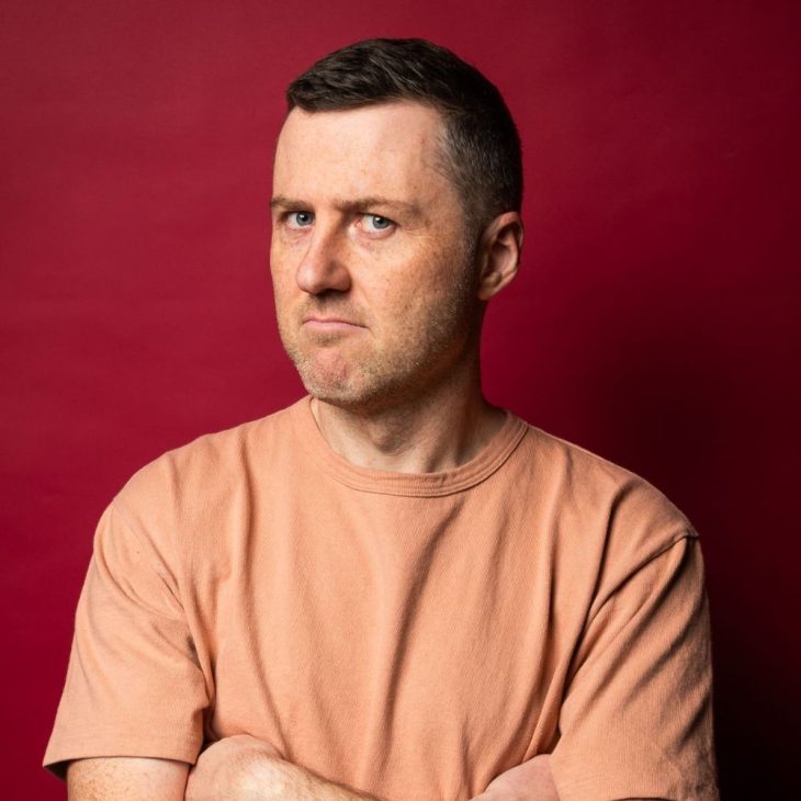 Lloyd’s head is turned to the side but his eyes are looking directly into the camera. He has his arms crossed and he has an annoyed expression on his face. He is wearing an orange tshirt against a red backdrop. He has short brown hair and blue eyes.