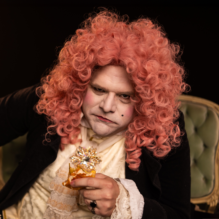 Image is of Greg Larsen dressed up in an 1800 period costume. He is wearing a pink permed wig, black jacket, and peach vest. He has 1800 period makeup and is sitting on a sage green velvet chaise. He is eating a cheeseburger.