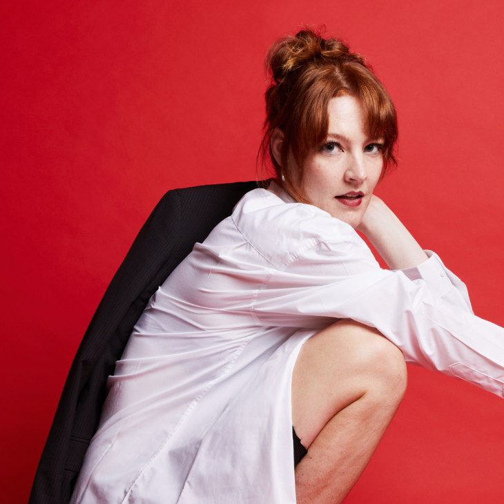 Image is of Lou Wall. Lou is crouching down to the right; their right arm is draped over their right knee and their left arm is holding a black jacket over their left shoulder. Their face is directed toward the camera. They have orange hair, red lips and are wearing a white button-down shirt with black shorts. They are in front of a red backdrop.