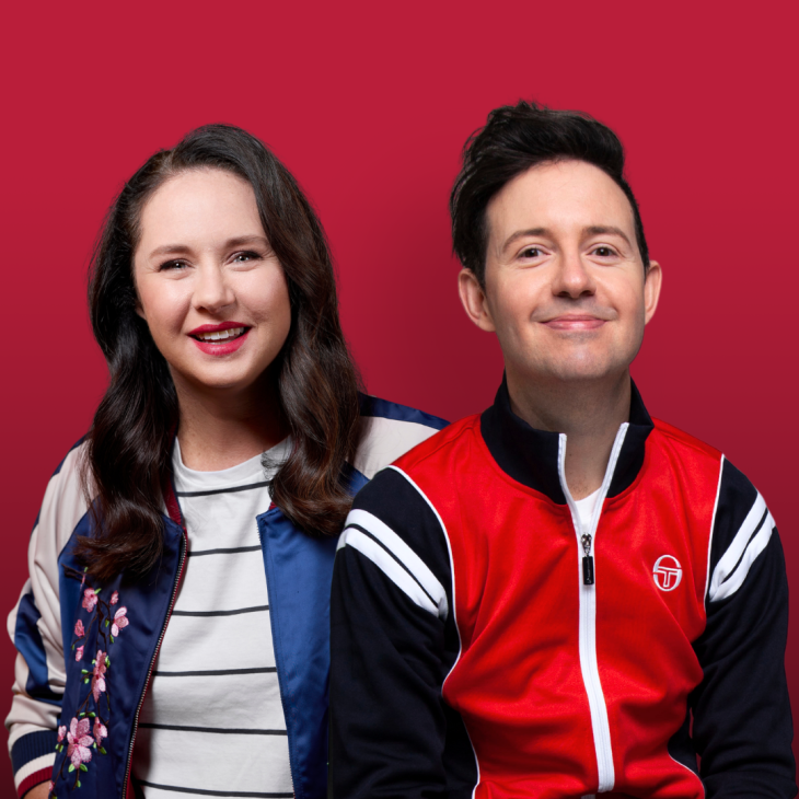 Mel Buttle sits behind and to the left of Nath Valvo. Mel has long brown wavy hair and red lipstick. She is wearing a blue jacket with a black and white stripy shirt. She is smiling and looking directly into the camera. Nath smiles at the camera with his head tilted back and up a little. His black mop of hair sits up on his head and his lips are pursed shut, like he might be about to laugh. He wears a track suit top – the body is red, the arms are black and it has white trim over the shoulders and down the zipper, which runs from top to bottom of the jacket. Tej background is maroon.