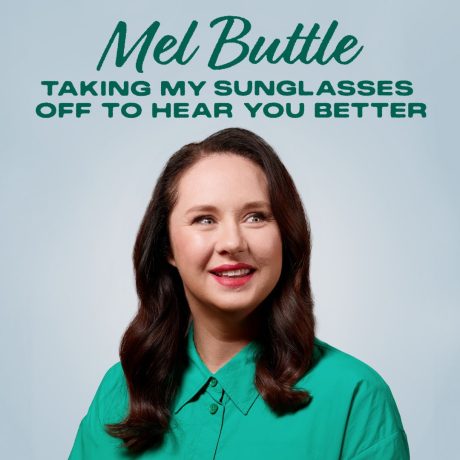 Mel Buttle smiling in front of a blue backdrop. She wears a teal shirt.