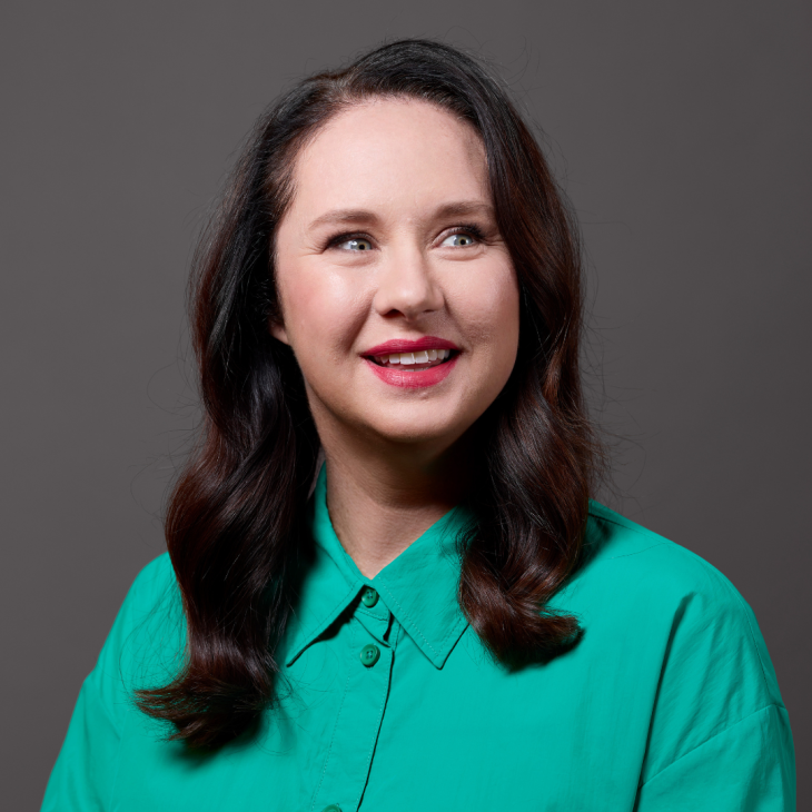 Mel Buttle smiling in front of a grey backdrop. She wears a teal shirt.
