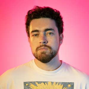 Image is of Nick White. Nick has short brown hair and a short beard. He is wearing a white t-shirt and is staring directly into the camera. He is standing in front of a pink background.
