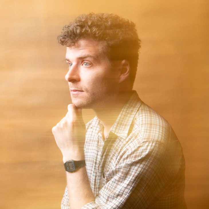 Oliver is standing on an angle giving a side profile. His hand is resting underneath his chin. He has short curly hair and blue eyes. He is wearing a brown checked shirt. The image has a blurry texture across the image.