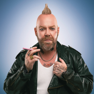 Image is of Pete Helliar. Pete is dressed as a biker with a blonde mohawk and tattoos. He is wearing a white singlet, black leather jacket, and gold jewellery. Pete is staring directly into a camera lens. He is holding a big boss pink cigar.