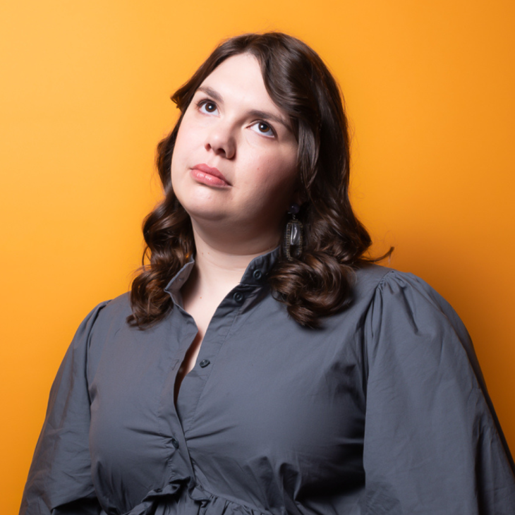 Prue Blake has short brown wavy hair. She is wearing a grey dress. She is looking up with a curious expression on her face. The background is orange.