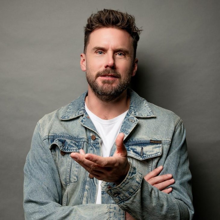 Dave is wearing a white shirt with a denim jacket. He is looking directly at the camera with his hand slightly raised. The background is grey.