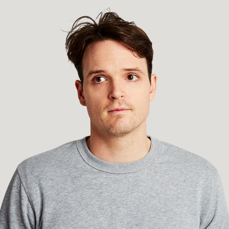 Tom Walker stands front on to the camera, with a neutral expression and his eyes looking off to his right. He has short dark brown hair, brown eyes and very short stubble. He wears a graymail t-shirt with small ribbing around the neck. The background is off white.