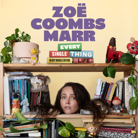 Image is of Zoe Coombs Marr. Her head is floating in a bookshelf. Her long dark hair is draped over the books and trinkets that cover the shelf.
