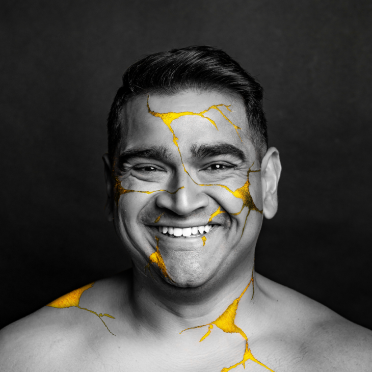 Image a black and white picture of Dilruk Jayasinha. Dilruk is looking directly into the camera and smiling with his teeth. Dilruk is shirtless and covered in gold cracks.