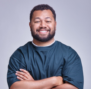 Image is of Joe Daymond. Joe is wearing a navy blue t-shirt and white pearl necklace. He has black short curly hair and a black beard. He is smiling directly into the camera with his arms crossed in front of his body.