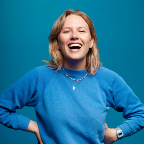 Image is of Jordan Barr. Jordan has short blonde hair, blue eyes and is wearing a blue pullover sweater with silver jewellery, she is looking into the camera and laughing. She is standing in front of a blue background.