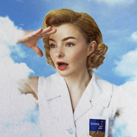 Image is of Abby Howells. Abby has short wavy blonde hair, blue eyes and bright red lips. She is wearing a white buttoned vest. She is standing amongst clouds, with her left arm raised above her eyes. She is peering off into the distance.