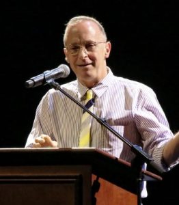 David Sedaris stands at a lectern. He smiles and is speaking into a microphone.